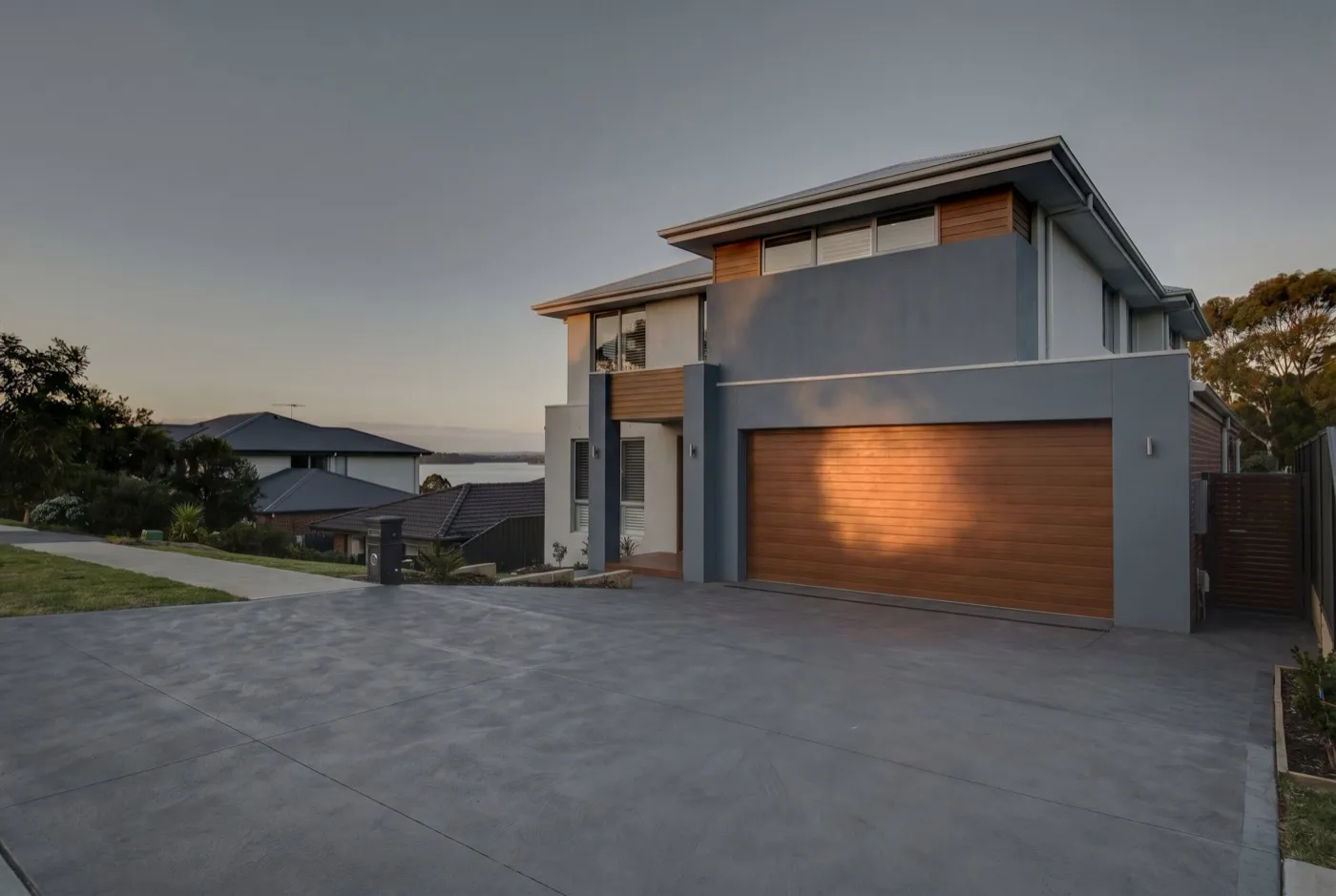A steep concrete driveway rising to a two-storey home with a wide timber-look sectional garage door, lake water visible between rooftops below